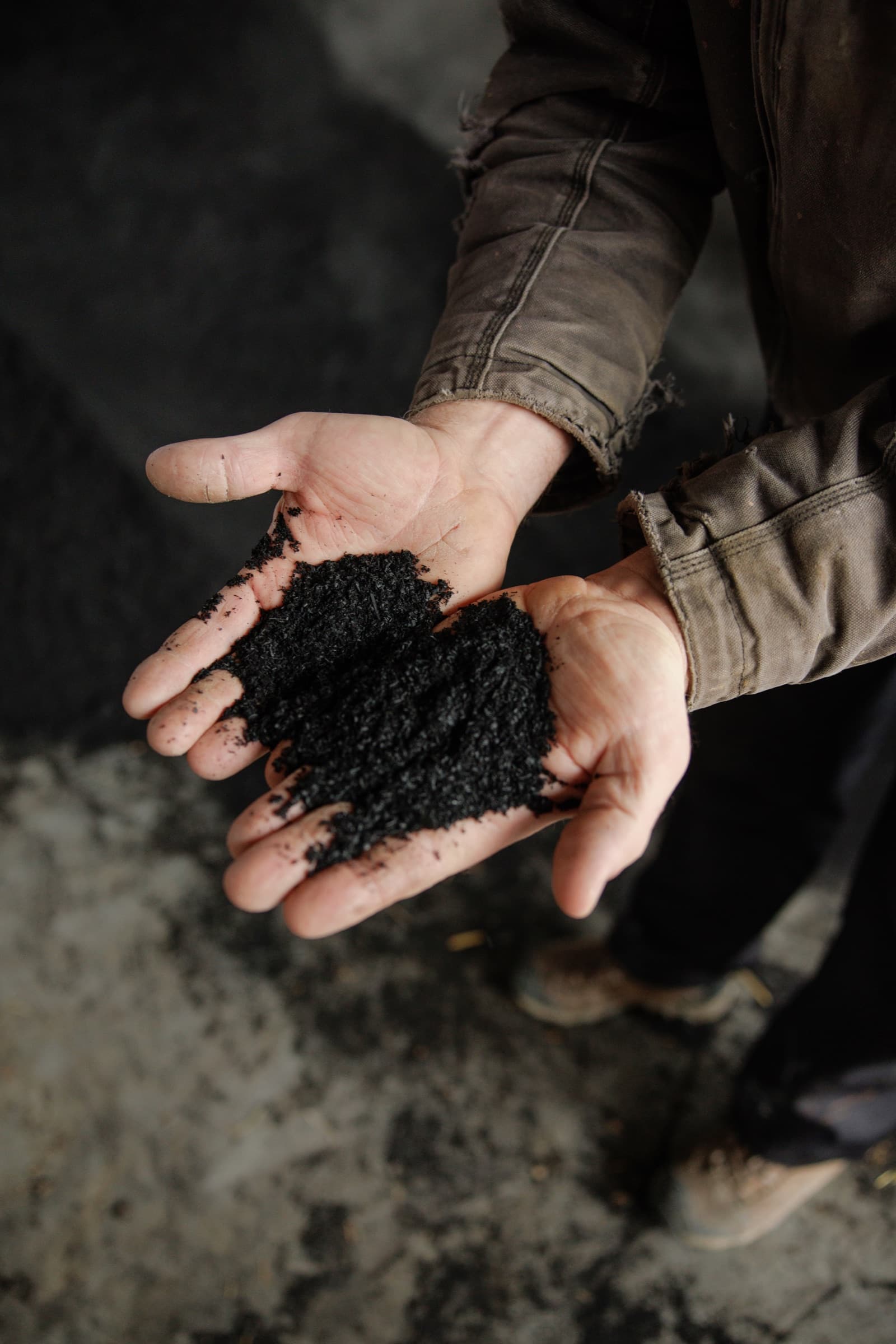 Two cupped hands hold a mound of AeroChar, a coarse black engineered biochar, on the floor of a barn.