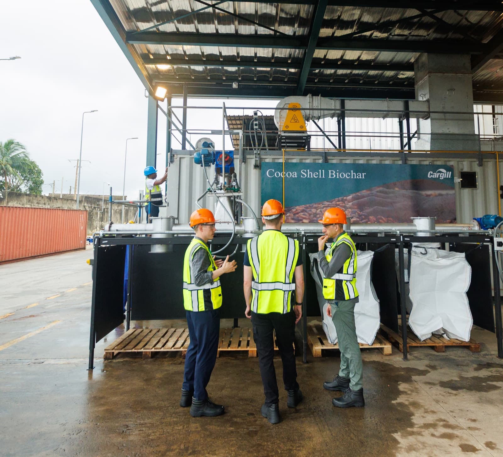 Three people in hi-vis vests in front of the containerized cocoa shell biochar unit in Côte d’Ivoire, supersacks filling under the canopy.