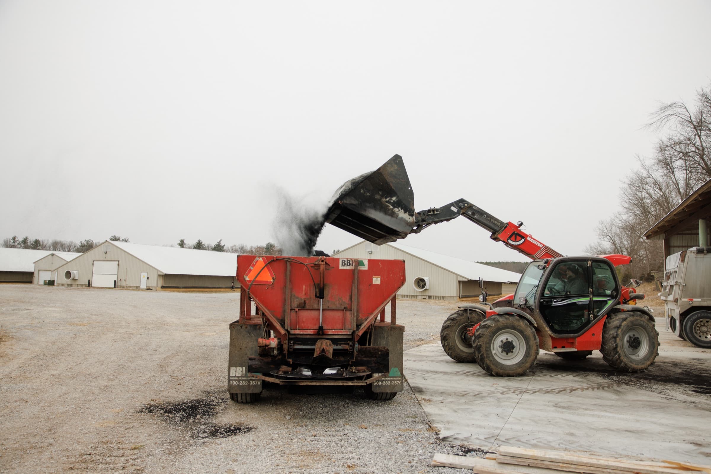 A telehandler tips a bucket of AeroChar into a BBI spreader in the yard, with the broiler houses of the complex behind.